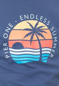 Ηλιοβασίλεμα με φοινικόδεντρο πάνω από μπλε κύματα, κείμενο «Pier One - Endless Summer» σε κυκλικό σχέδιο σε σκοτεινό υφασμάτινο φόντο.