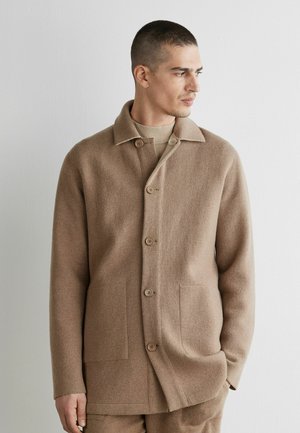 Man med kort hår som bär en knäppt ljusbrun ullkavaj och matchande byxor, tittar åt sidan mot en enkel vit bakgrund.