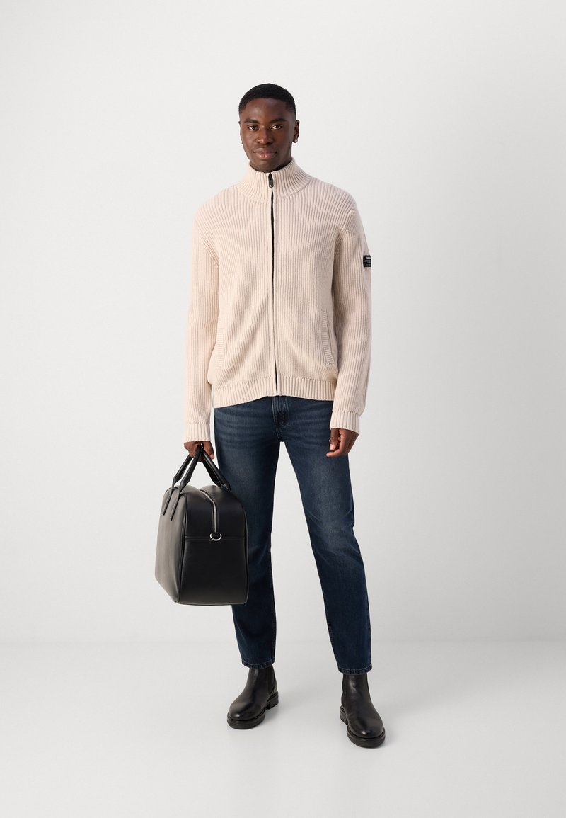 Jasno beżowy sweter z prążkowaną teksturą i zamkiem błyskawicznym, z wysokim kołnierzem, w połączeniu z ciemnymi dżinsami i czarnymi skórzanymi botkami, trzymający czarną skórzaną torbę.