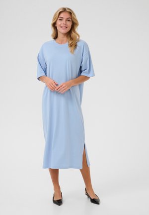 Robe midi bleu clair en tissu doux, avec des manches courtes, un col rond et des fentes latérales pour plus de liberté de mouvement. Escarpins noirs portés.