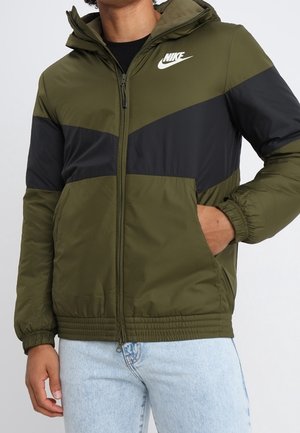 Mann trägt olivgrüne und schwarze Nike Kapuzenjacke mit Reißverschluss, die Hände in den Taschen, kombiniert mit hellblauen Jeans.