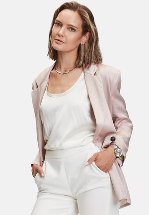 Lyserød satin blazer over en hvid tanktop, parret med hvide bukser. Sølv halskæde og ur, lange øreringe, glat tekstur.