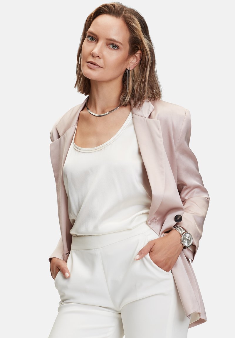 Blazer in satin rosa chiaro sopra una canotta bianca, abbinato a pantaloni bianchi. Collana e orologio argentati, orecchini lunghi, texture liscia.