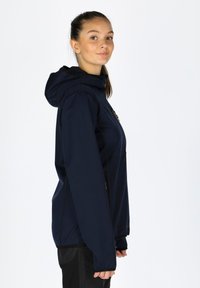 Marineblaue Softshell-Jacke mit Kapuze, ausgestattet mit einem Reißverschluss vorne, elastischen Bündchen und einem leicht taillierten Design. Glatte Textur, minimale Details.