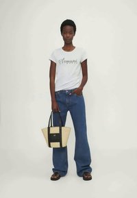 Bílé tričko s šedým textem, modré rozšířené džíny a béžová taška s černými akcenty. Modelka ve černých sandálech stojící na světlém pozadí.