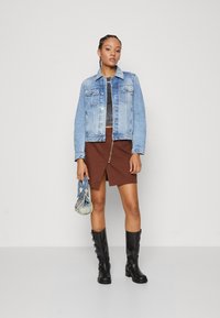 Veste en denim bleu clair avec des poches, haut gris, jupe marron avec une fermeture éclair, et bottes noires montantes avec un matériel décoratif.
