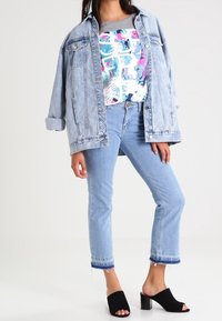 Chaqueta de mezclilla azul claro sobre una camiseta gráfica colorida. Jeans cortos azul claro con dobladillo deshilachado y tacones de bloque abiertos negros.