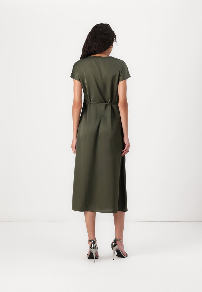 Vestido midi verde olivo con mangas cortas, de textura suave y detalle de cintura atada. Llevado con zapatos de tacón plateados, mostrado desde atrás.