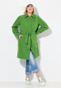 Cappotto trench verde con cintura, maniche lunghe, tasche laterali e colletto a punte; abbinato a jeans azzurri con risvolti e sneaker bianche.