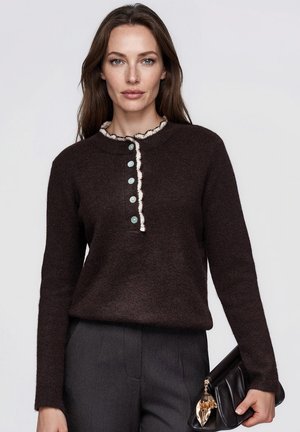 Une femme porte un pull marron foncé avec des bordures et des boutons couleur crème, un pantalon gris et un sac à main noir, sur un fond uni.