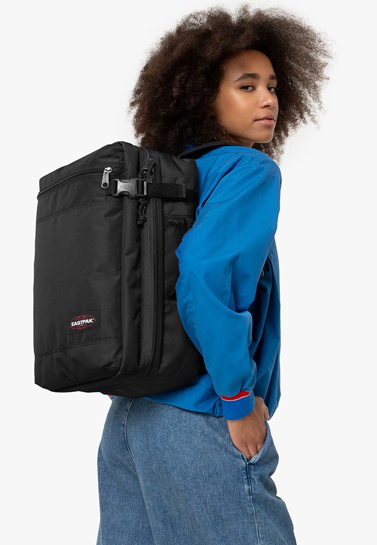 Black backpack with textured fabric, multiple zippers, and a front patch logo, worn over a blue jacket with red accents and denim jeans.
