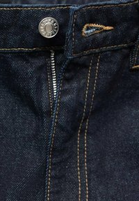 Gros plan sur la zone de la fermeture éclair d'un jean denim bleu foncé avec un bouton argenté et des coutures orange contrastantes.