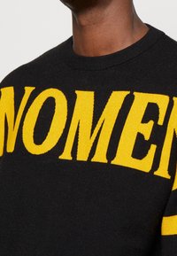 Pull noir avec un texte jaune vif « WOMEN » en lettres capitales sur le devant. Tissu en maille avec un col rond et un col côtelé, présentant une texture lisse.