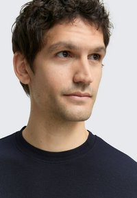 Granatowy, teksturowany sweter z okrągłym dekoltem i prążkowanym kołnierzem. Krótkie jasnobrązowe włosy, minimalne owłosienie na twarzy i neutralny wyraz twarzy.