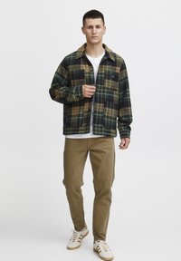 Camicia di flanella a quadrati verdi e marroni, con bottoni frontali, colletto a punta e taschino. Abbinata a pantaloni khaki e sneakers bianche.