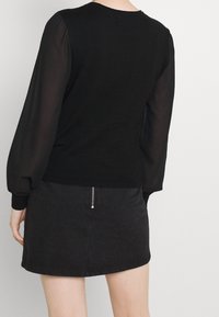 Svart tröja med genomskinliga långa ärmar, rund halsringning och ribbad fåll, kombinerad med en mörk denimkjol med synlig dragkedja.