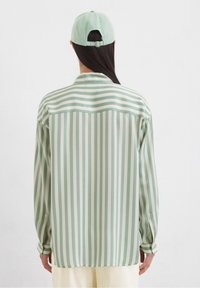 Camicia a righe con strisce verticali verde menta e bianco sporco, maniche lunghe, yoke posteriore e vestibilità rilassata; abbinata a un cappellino coordinato.