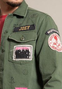 Närbild av en grön jacka med lappar som läser "JOEY," "BEERBONES," och "ROCK'N'ROLL RENDEZVOUS," bärs över en röd skjorta.
