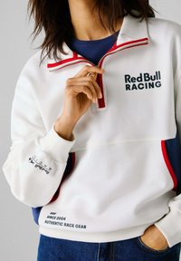 Biely fleece polovičný zips s červenými akcentmi a námorníckymi bočnými panelmi. Obsahuje logo "Red Bull Racing" a ďalší text na rukáve.