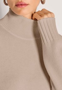 Nahaufnahme einer Frau, die einen beigen gerippten Rollkragenpullover trägt, den Kragen mit einer Hand hält und große Creolen trägt.