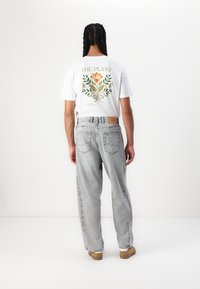 Tricou alb cu grafică florală în nuanțe de portocalie și verde pe spate, asociat cu blugi gri deschis, largi și încălțăminte bej.