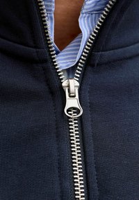 Tumesinine tõmblukuga jakk, millel on sile metallist tõmblukk ja pehme tekstuur. All on sinise ja valge triibuline kaelusega särk.