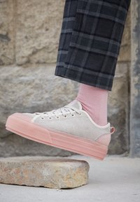 Beige kanvas-sneakers med en texturerad yta och en rosa gummisula. Har vita snörningar och en vadderad krage, tillsammans med ljusrosa strumpor.