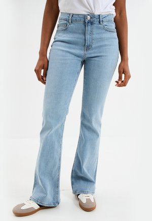 Person trägt hellblaue High-Waist Flared Jeans mit einem weißen, hineingesteckten Oberteil und weißen sowie grauen Turnschuhen, steht vor einem weißen Hintergrund.