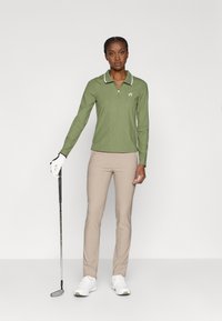 Zaļš garām piedurknēm golfa polos ar baltu apkakli un priekšējo rāvējslēdzēju, savienots ar bēšiem golfa biksēm un baltām kedām, tur rokā golfa nūju.