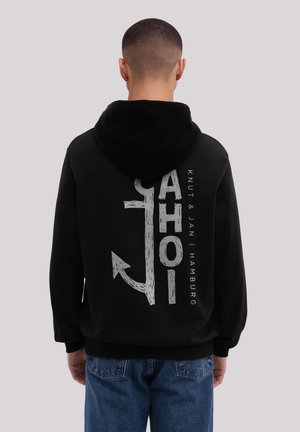 Person mit schwarzem Hoodie, auf dem ein großes weißes Anker-Graphic und der Text "AHOI" auf dem Rücken zu sehen sind, kombiniert mit blauen Jeans, steht vor einem grauen Hintergrund.