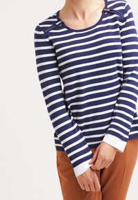Navy and white striped long-sleeve top with a round neckline, featuring shoulder zippers and white cuffs. Worn with brown pants.