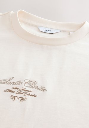 Kremowy t-shirt z prążkowanym dekoltem i brązowym haftowanym napisem „Santa Clarita” oraz ozdobnym akcentem.