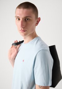 Tricou cu mânecă scurtă, de culoare albastră deschisă, cu un logo roșu pe piept, asociat cu o geantă neagră, purtată de o persoană cu părul scurt.