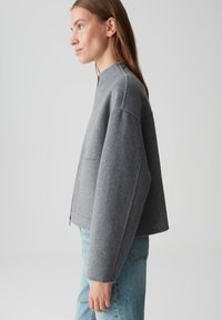 Vrouw in een grijze woljas met rits en lichtblauwe spijkerbroek, staand in profiel tegen een effen lichtgrijze achtergrond.