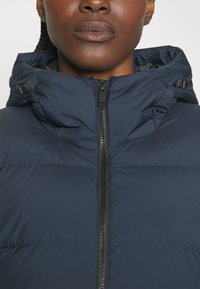 Jūras spēku pufersjakets ar rāvējslēdzēju priekšpusē, augstu apkakli un regulējamu kapuci. Gluda, quilted auduma struktūra ar minimālu aparatūras detaļu, lai nodrošinātu siltumu.