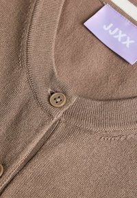 Ανοιχτό καφέ πλεκτή ζακέτα με στρογγυλή λαιμόκοψη, κλείσιμο με κουμπιά και ορατή λιλά ετικέτα ρούχου με την επιγραφή "JJXX".
