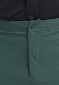 Gros plan sur un pantalon vert foncé avec un bouton-pression recouvert de tissu et une fermeture à glissière cousue, porté avec un haut noir.