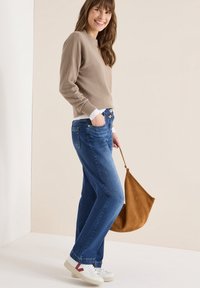 Béžový žebrovaný svetr, modré džíny rovného střihu, bílé tenisky s červenými detaily a hnědá semišová kabelka přes rameno, stojící na světlém pozadí.