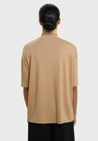Beige kortærmet top med høj hals, glat tekstur, løs pasform og rund kant, set bagfra.