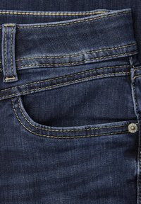 Kieszeń ciemnoniebieskich dżinsów z widoczną pętlą na pasek, żółtym przeszyciem i metalowym nitem po prawej stronie.