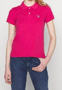 Frau trägt ein leuchtend pinkes Poloshirt mit kurzen Ärmeln und einem kleinen gestickten Vogel-Logo, kombiniert mit dunkelblauen Jeans.