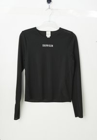 Zwarte langemouwen mesh shirt met "CALVIN KLEIN" in witte letters over de borst gedrukt, hangend aan een heldere plastic hanger tegen een witte achtergrond.