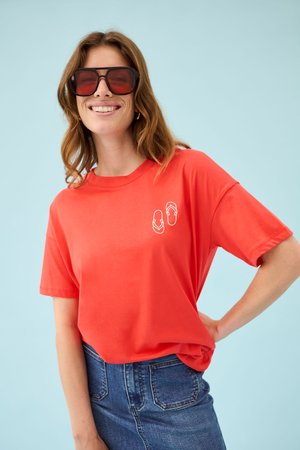 Femme portant de grandes lunettes de soleil rouges, un t-shirt rouge vif avec des icônes de tongs blanches et un jean bleu taille haute sur fond bleu clair.