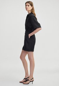 Combinaison noire à manches courtes avec col, taille élastique et deux poches latérales. Portée avec des sandales noires à lanières ornées d'accents dorés.