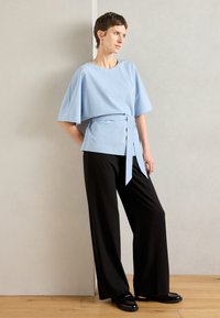 Ανοιχτό μπλε κοντομάνικο μπλουζάκι με δέσιμο στη μέση, συνδυασμένο με μαύρες παντελόνες με ψηλή μέση και φλατ μαύρες loafers. Χαλαρές υφές.