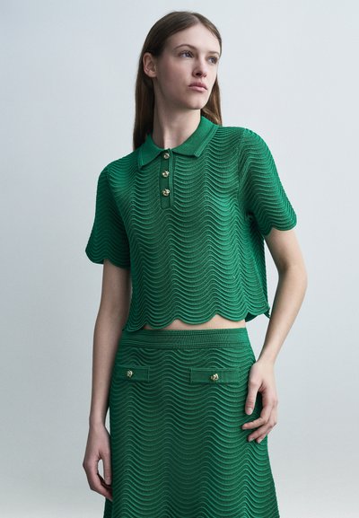 Femme portant un haut polo en tricot vert à manches courtes et une jupe assortie avec un motif texturé ondulé et des détails de boutons dorés.