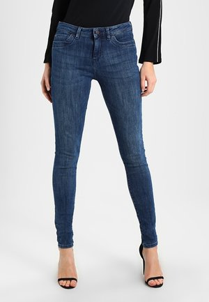 Vrouw die donkerblauwe skinny jeans en zwarte sandalen met hoge hakken draagt, staand tegen een effen lichte achtergrond.