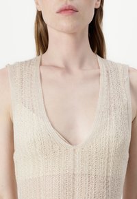 Bež pleteni top brez rokavov z globokim V-izrezom, ki ima strukturiran odprt vzorec in nekoliko širše naramnice.