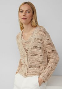Strickjacke in hellem Beige mit Lochmuster, ausgestattet mit einem V-Ausschnitt, zwei großen Knöpfen und gerippten Bündchen, getragen über einem passenden Tanktop.