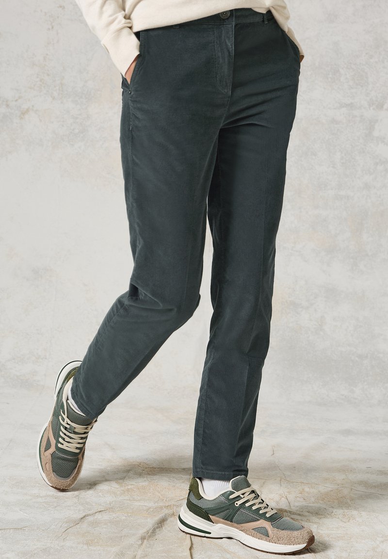 Ciemnozielone spodnie z kordurowej tkaniny o gładkiej fakturze, prostym kroju i bocznymi kieszeniami, połączone z zielono-beżowymi butami sportowymi.